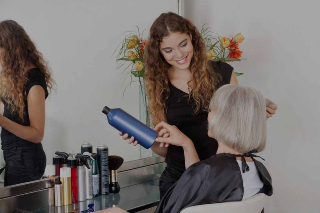 Uma jovem cabeleireira sorridente de cabelo encaracolado segura um frasco azul de produto capilar enquanto aconselha uma mulher idosa de cabelo grisalho sentada numa cadeira de salão.