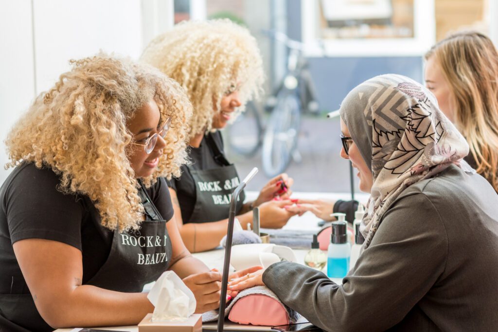 Duas manicures de cabelo loiro encaracolado com aventais pretos onde se lê "Rock & Roll Beauty" fazem a manicure a clientes numa mesa comprida. Uma cliente usa um hijab cinzento.