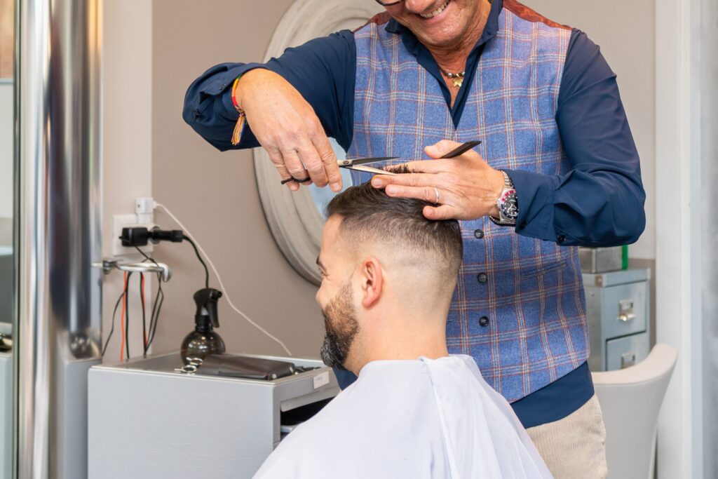 Um cabeleireiro com um colete de xadrez azul corta o cabelo de um cliente com tesoura e pente num salão. O cliente tem cabelo curto, barba e usa uma capa de corte branca.