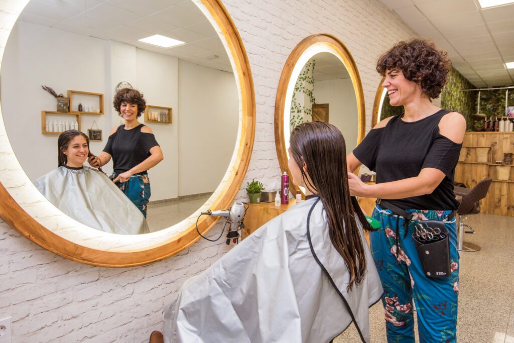 Una peluquera sonriente con pelo corto rizado cepilla el cabello largo y húmedo de una cliente frente a un gran espejo redondo iluminado. La escena se refleja en el espejo, mostrando el interior de la peluquería.