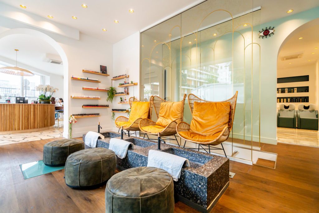 Interior elegante e luminoso de um salão de beleza que dispõe de uma zona de pedicure com três poltronas de verga amarelo-mostarda, lavatórios de pedra com toalhas brancas e pufs cinzentos a condizer sobre um piso de madeira.