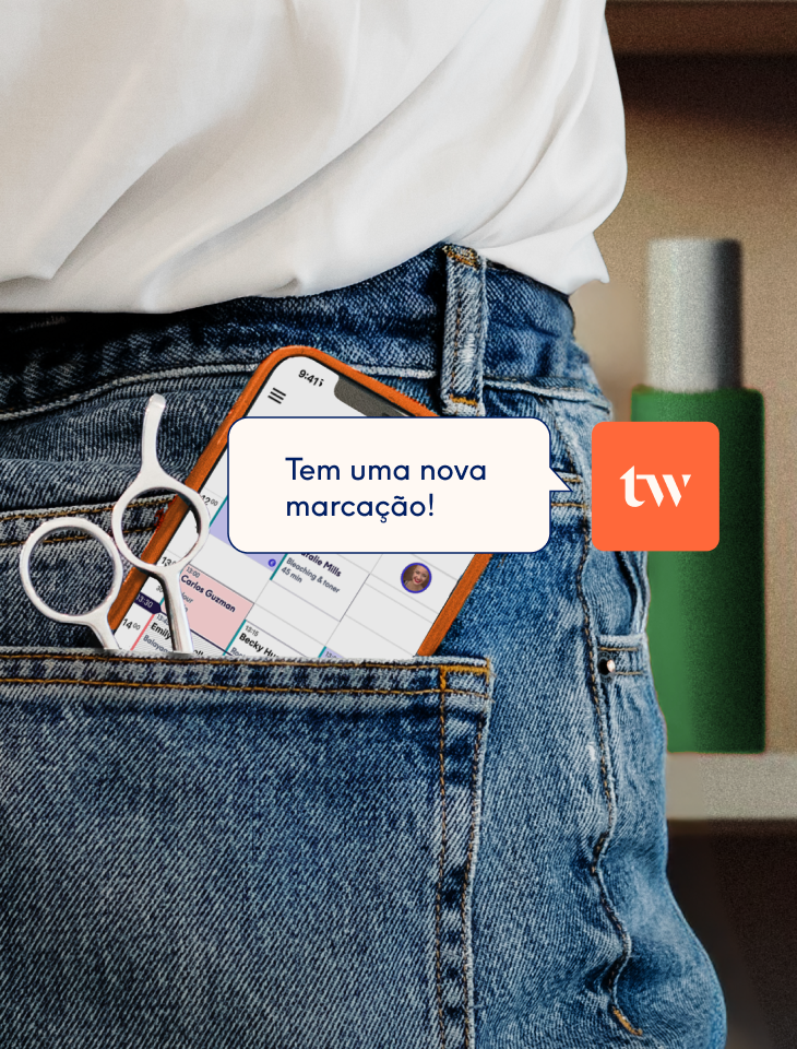 Um smartphone e uma tesoura de cabeleireiro no bolso de trás de umas calças de ganga, com o ecrã a mostrar a agenda de marcações da app Treatwell e uma notificação de "Tem uma nova marcação".